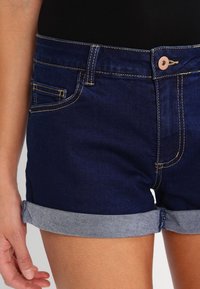 Close-up van een persoon die donkerblauwe denim shorts met omgezoomde zoom, contrasterende stiksels en een knoopsluiting draagt, staand tegen een effen achtergrond.