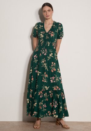 Kobieta w długiej sukience maxi w ciemnozielony kwiatowy wzór, z krótkimi rękawami i dekoltem w serek, stoi na tle jasnej, jednolitej ściany z rękami za plecami.