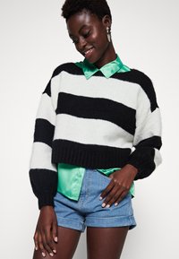 Svart-og-hvitt-stripet avskåret genser med ribbede mansjetter, kombinert med en grønn satengskjorte med krage og denimshorts. Glatt tekstur gjennom hele.