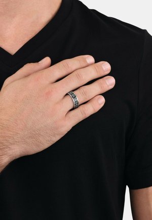 Schwarzes kurzärmliges V-Ausschnitt-Shirt mit einer Hand, die einen strukturierten Metallring mit komplizierten Mustern in Silber und Schwarz hält.