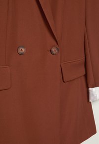 Veste croisée marron à deux boutons, poches à rabat, avec une manche de chemise blanche retroussée visible sur le côté droit.