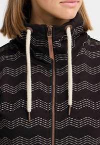 Giacca nera con zip e colletto alto, caratterizzata da un motivo zigzag bianco. Dettagli includono lacci color crema e hardware in bronzo.