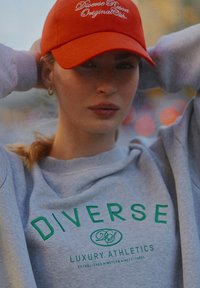 Szara bluza z zielonym napisem "DIVERSE LUXURY ATHLETICS", w połączeniu z jasnopomarańczową czapką z białym haftowanym napisem.