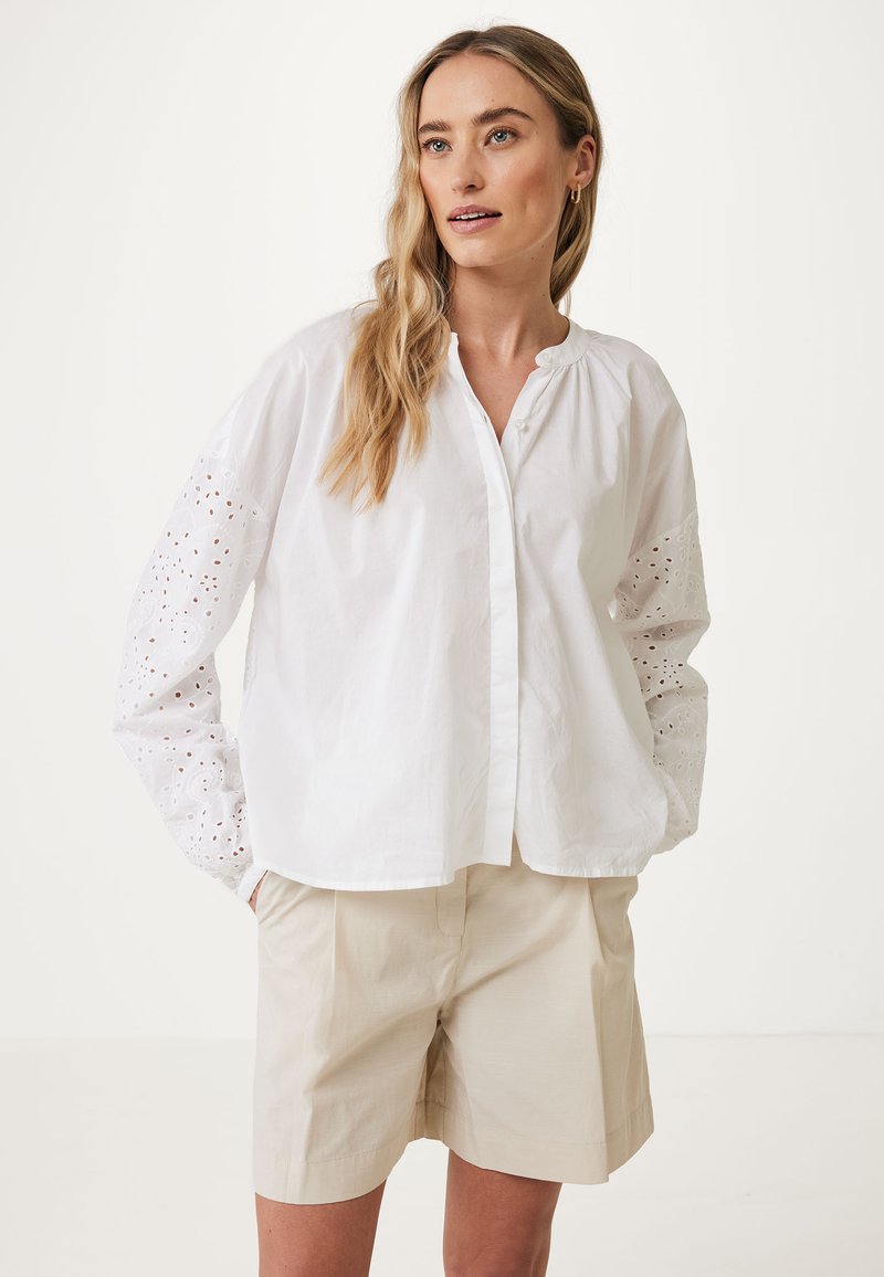 Vit bomullsblus med broderade ögonskydd på ärmarna, knappstängning fram och rund halsringning. Matchad med beige shorts.