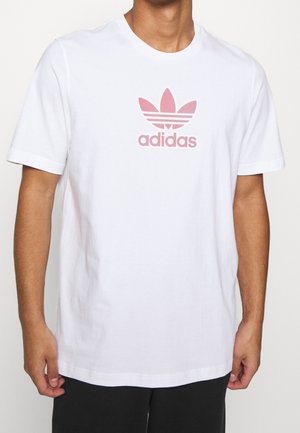 Biała bawełniana koszulka z różowym logo Adidas i grafiką koniczyny na przodzie. Krótkie rękawy i okrągły dekolt.