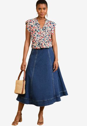 Bloemenprint top met gerimpelde mouwen, gecombineerd met een lange, donkere spijkerrok. Model houdt een geweven tas vast en draagt tan kleurige sandalen met bandjes.