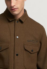 Giacca di lana marrone con colletto ripiegabile, bottoni a pressione neri e due tasche sul petto con dettagli del logo. Texture liscia.