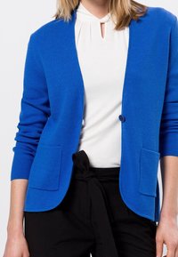 Blazer de punto azul con dos bolsillos frontales y cierre de un botón. Lleva encima de una blusa blanca, con escote redondo y pantalones negros.