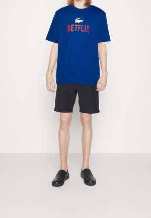 Camiseta azul de cuello redondo con texto rojo "NETFLIX" y un logo de cocodrilo blanco, combinada con pantalones cortos negros y zapatillas negras.
