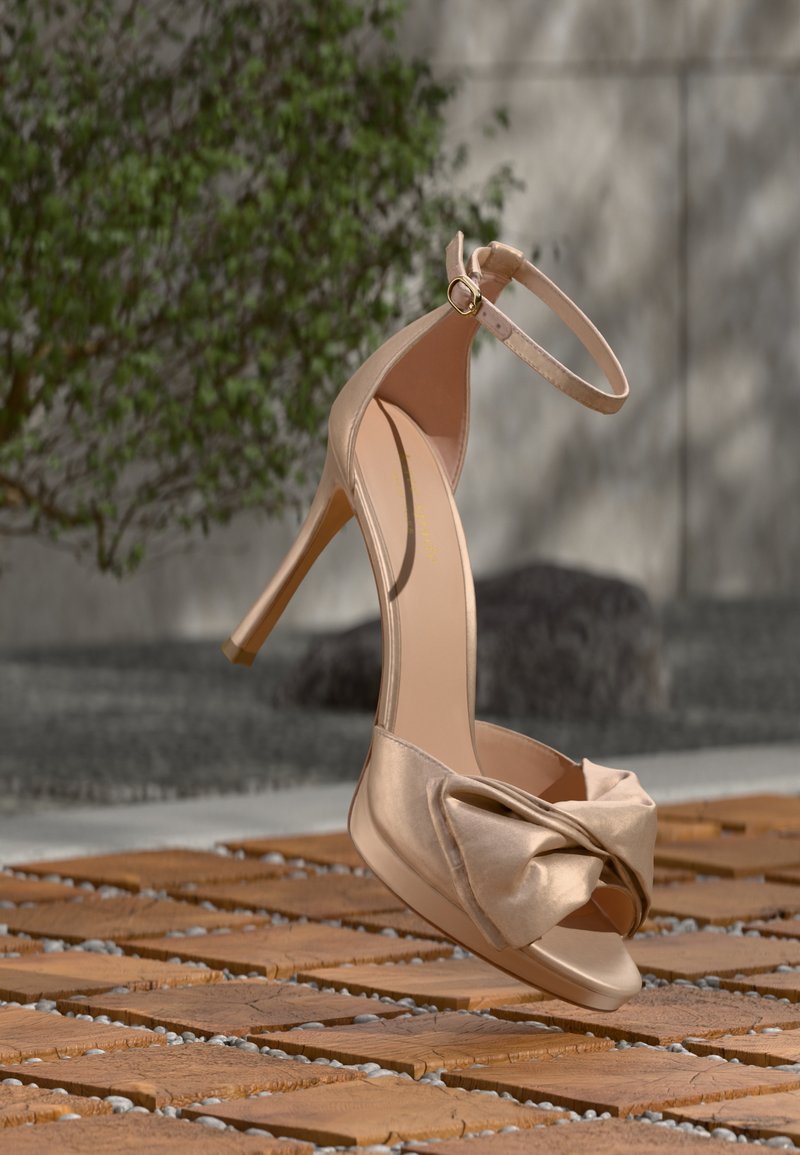 Μπεζ ψηλοτάκουνο σανδάλι με λουράκι στον αστράγαλο και μεγάλο φιόγκο στο δάχτυλο, τοποθετημένο όρθιο σε ξύλινο και βοτσαλωτό πάτωμα σε εξ ωτερικό χώρο.