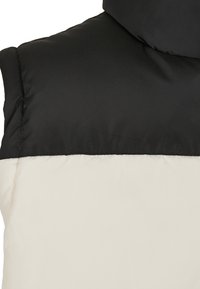 Gepolsterte Weste mit einem schwarzen oberen Bereich und einem cremefarbenen unteren Bereich. Glatte Textur mit kontrastierenden Farben; runder Halsausschnitt.