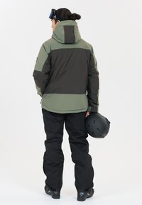 Schneejacke mit einem dunkelgrünen und schwarzen Farbblockdesign, aus strapazierfähigem Stoff, mit Kapuze und Reißverschlusstaschen. Kombiniert mit schwarzen Hosen.