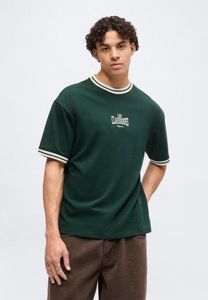 Jeune homme aux cheveux bouclés portant un t-shirt vert foncé avec un col et des poignets rayés blancs, et un pantalon marron, debout devant un fond uni.