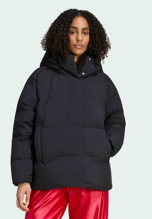 Kvinde med krøllet hår iført en sort dunjakke med hætte og røde Adidas træningsbukser, stående mod en lys baggrund.