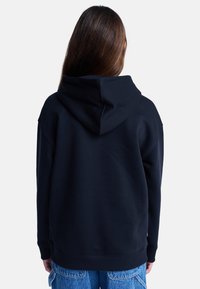 Felpa nera con cappuccio, dal taglio morbido e polsini a coste, con un grande cappuccio e dettagli sulla schiena. Indossata con jeans blu, dal design minimalista.