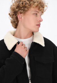 Giacca nera con zip e colletto in finta shearling bianca. Presenta due tasche frontali e una superficie liscia. Indossata sopra una camicia chiara.