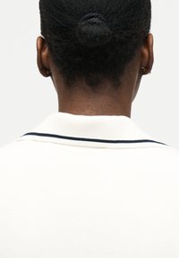 Personne à la peau foncée et aux cheveux courts naturels attachés en petit chignon, portant un haut blanc avec un col rayé noir, vue de dos.