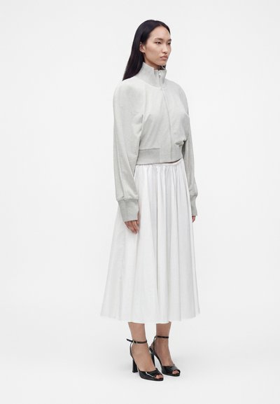 Femme portant une veste zippée gris clair, une jupe midi plissée blanche et des talons ouverts noirs avec des brides à la cheville, debout devant un fond blanc.