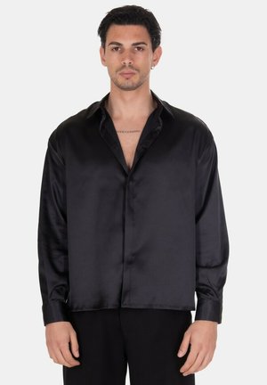 Uomo che indossa una camicia nera di raso a maniche lunghe e collo aperto, in piedi davanti a uno sfondo chiaro uniforme, rivolto in avanti.
