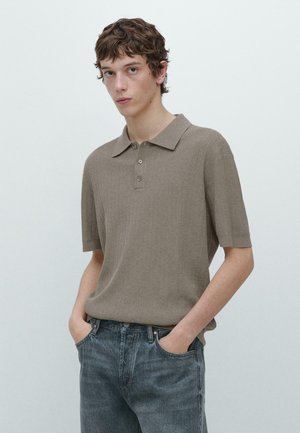 Giovane uomo con capelli corti e ricci indossa una polo a maniche corte in tessuto lavorato color taupe e jeans blu, in piedi con le mani in tasca davanti a uno sfondo neutro.