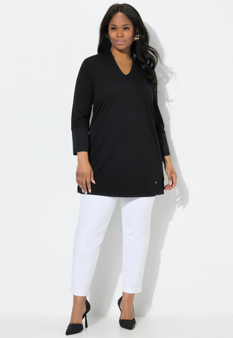 Schwarze Longsleeve-Tunika mit V-Ausschnitt und seitlichen Schlitzen, kombiniert mit weißen, figurbetonten Hosen und schwarzen, spitzen Absätzen. Goldene Akzentdetails vorhanden.