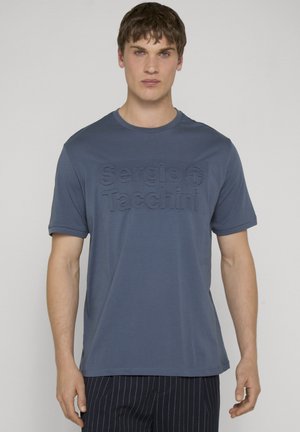 Giovane uomo che indossa una t-shirt blu Sergio Tacchini e pantaloni scuri a righe sottili, in piedi davanti a uno sfondo chiaro e uniforme.