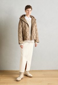 Veste matelassée marron avec capuche, un pull en tricot beige en dessous, une jupe crème avec une fente sur le côté, et des bottes beiges à lacets sur un sol en bois.