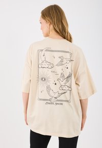 Beige oversized t-shirt met zwarte grafische ontwerpen van vogels, wolken, een maan en de tekst "Positive Sincere" op de achterkant.