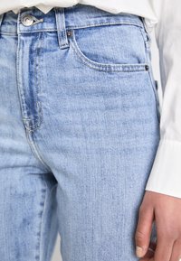 Närbild av ljusblå denimjeans i midjan och övre låret, bärs tillsammans med en vit långärmad skjorta, som visar metalldetaljer på knappar och fickor.