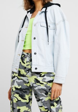 Kvinde iført lys denimjakke med sort hætte over limegrøn crop top og gul-grøn camouflage cargobukser, stående med afslappet hånd.