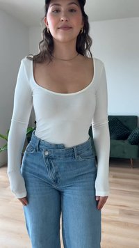 Weißes, langärmeliges, tailliertes Oberteil mit weitem Halsausschnitt, kombiniert mit hoch taillierten hellblauen Jeans. Das Outfit ist einfach und lässig.