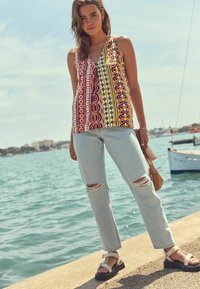 Haut sans manches à motifs colorés avec un décolleté en V, associé à un jean clair délavé avec des déchirures aux genoux, et des sandales blanches avec des semelles noires.
