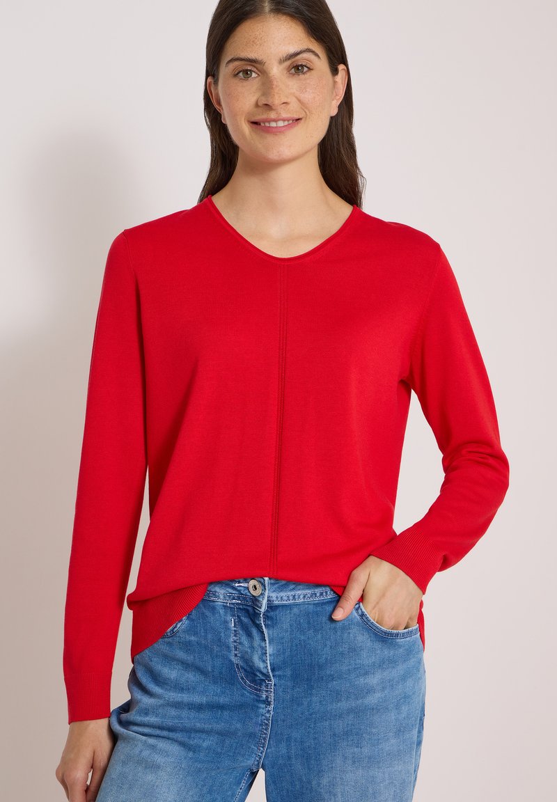 Femme portant un pull rouge à manches longues et un jean bleu, la main droite dans la poche, souriant à la caméra.