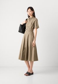 Khaki button-up shirt dress with short sleeves, fitted waist, and flared skirt. Paired with black open-toe mules and a black handbag.