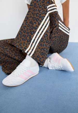 Person iført brune leopardmønstrede bukser med hvide striber og hvide sneakers med lyserøde striber, der sidder på en blå tæppebelagt gulv.