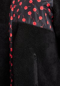 Schwarze Fleecejacke mit glänzendem Stoffpaneel und rotem Kirschmuster, akzentuiert durch eine Reißverschlusstasche an der Unterseite.