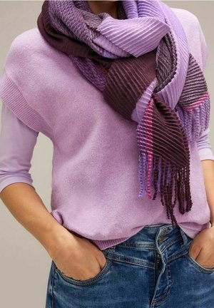 Personne portant un pull lavande avec les manches retroussées, un jean bleu et une écharpe texturée rayée violette enroulée autour du cou, les mains dans les poches.