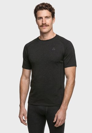 Mann mit braunen lockigen Haaren und Schnurrbart, der ein enganliegendes schwarzes Kurzarmshirt und schwarze Hosen vor einem schlichten grauen Hintergrund trägt.