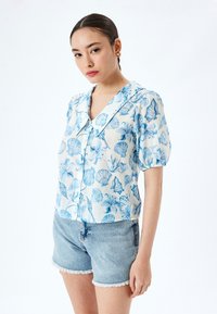 Kurzärmlige Bluse aus leichtem Stoff mit einem blauen Muschel- und Blumenmuster, ausgestattet mit einem eingeschnittenen Kragen und einem lockeren Schnitt. Dazu eine kurze Jeanshose.