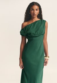 Grünes Satin-Ein-Schulter-Kleid mit drapiertem Oberteil, figurbetonter Silhouette und glatter Textur. Mit einem Armreif am einen Handgelenk.