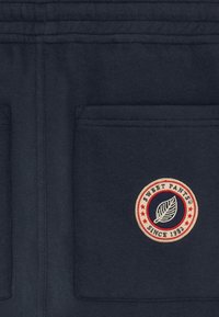 Pantalons de survêtement bleu marine avec deux poches au dos. Présente un logo rond avec une feuille et "SWEET PANTS® SINCE 1982" en accents crème et rouge.