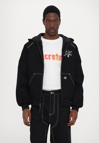 Black hooded jacket with white stitching, featuring a graphic on chest. Worn over a white shirt with vibrant pink lettering and black pants.
