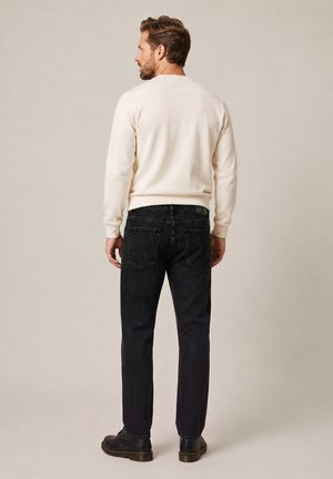 Homme aux cheveux courts et bruns portant une chemise crème à manches longues, un jean noir et des chaussures noires, debout de dos sur un fond neutre.