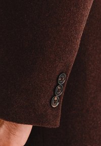Brązowa wełniana marynarka z mankietami przedstawiająca cztery czarne guziki z kontrastującą nicią, prezentująca teksturowany materiał i nowoczesny krój.