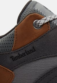 Primo piano di una sneaker Timberland grigia e marrone con dettagli in tessuto, suede, pelle, occhielli in metallo e logo Timberland in rilievo nero.