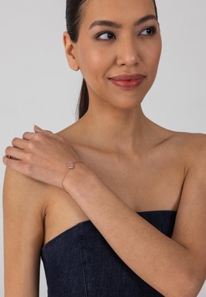 Frau in einem trägerlosen dunklen Oberteil trägt ein zartes roségoldenes Armband mit einem rosa Blumenanhänger am linken Handgelenk und hält ihre rechte Schulter.