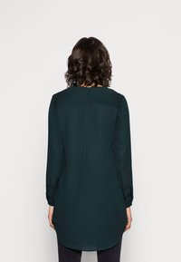 Mörkgrön tunika i slätt tyg, med långa ärmar, rund halsringning och en något rundad fåll. Inkluderar detaljer med en veck bak.