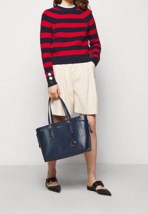 Granatowa skórzana torba typu tote ze złotymi okuciami, podwójnymi uchwytami i detalem w postaci frędzla. Dopełnieniem stroju jest sweter w czerwono-granatowe paski oraz beżowe szorty.