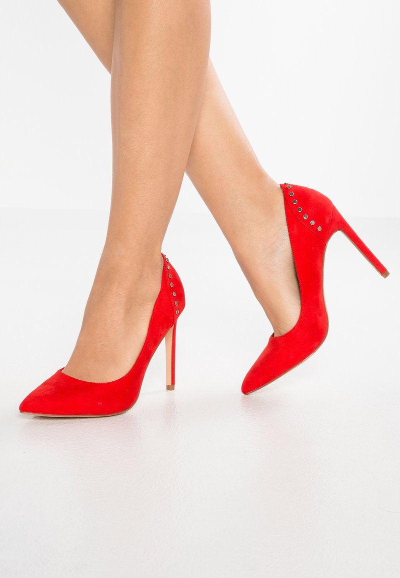 Escarpins en suède rouge avec un bout pointu, ornés de clous décoratifs le long du talon, et un design stiletto. Texture lisse et couleur vive.