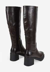 Botas de cuero marrón hasta la rodilla con una textura suave, tacón cuadrado y detalle de cremallera lateral. El diseño presenta una silueta simple y elegante.